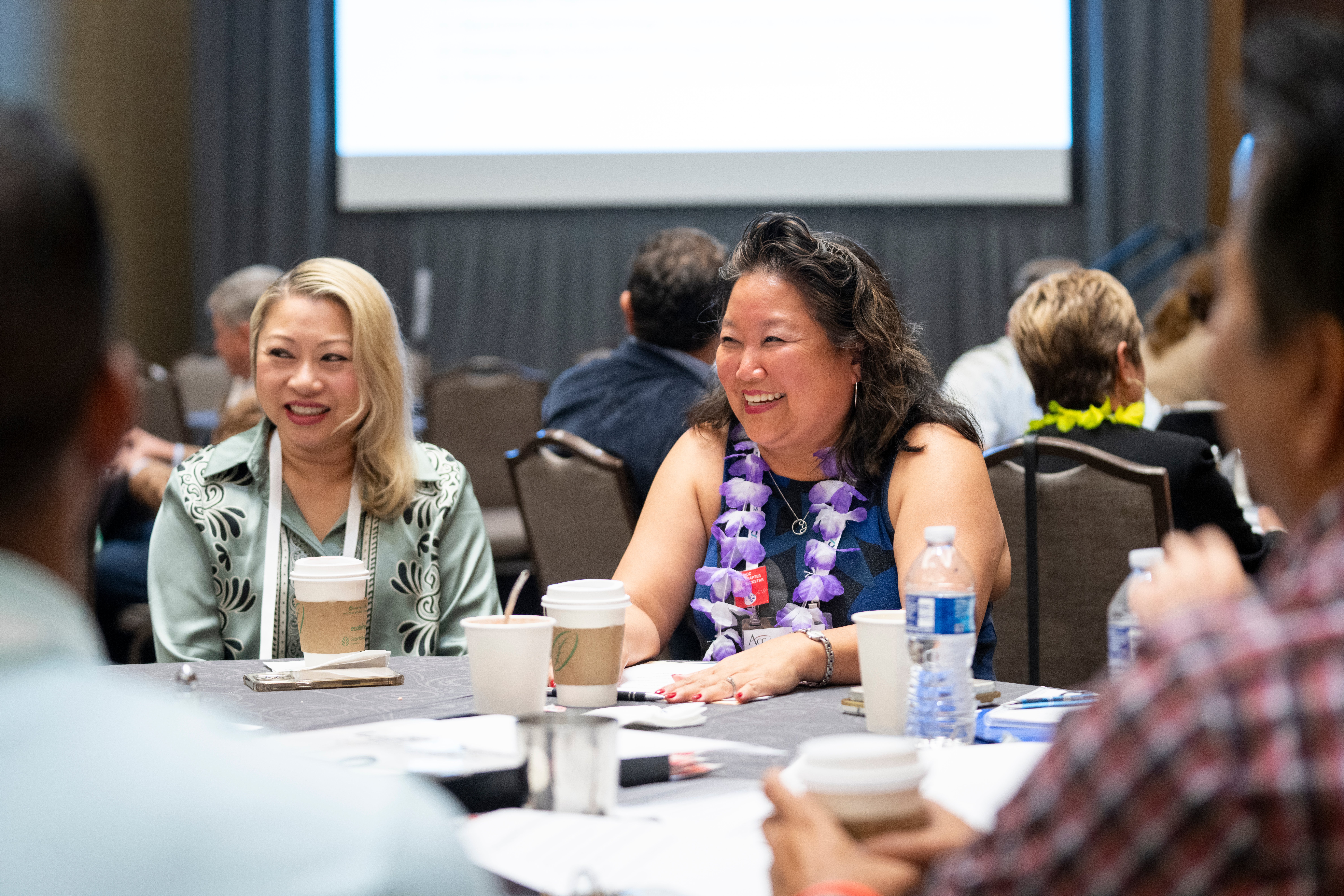 two people seated at table with cups of coffee near them, one person wearing a lanyard and the other person wearing a lei, listening to a speaker at the table and smiling